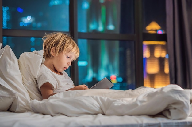 The boy uses the tablet in his bed before going to sleep on a background of a night city Children and technology concepts