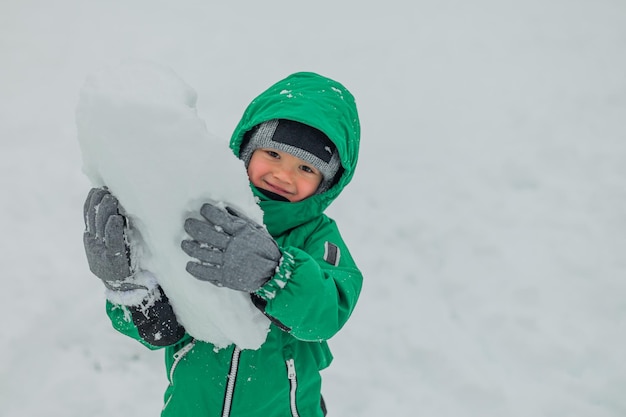 A boy walks outside in the snow the boy carries a large snowball in his hands the boy plays in the snow