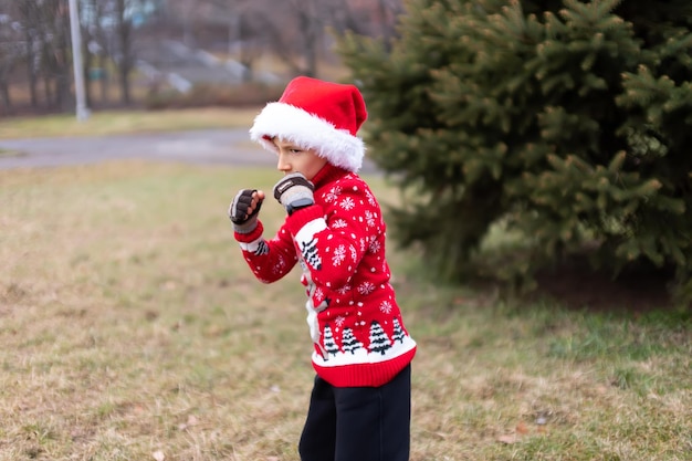 A boy in a warm Christmas sweater with a New Year's deer and a Santa hat on the background of a Christmas tree clenched his fists in front of him and stood in a fighting stance