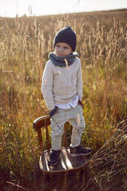 Boy in warm clothes stand on chair along a path on a field with dried grass