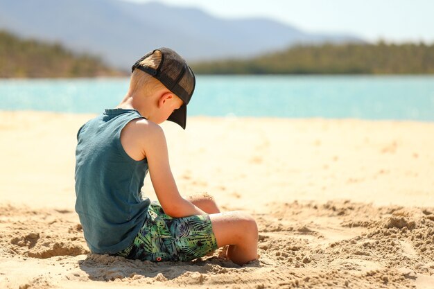 Boy wearing a cap sitting at the beach playing in the sand