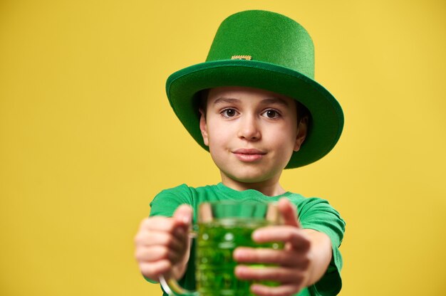 A boy wearing a green leprechaun hat holds a glass of green\
drink and holds it out in front of him to the camera