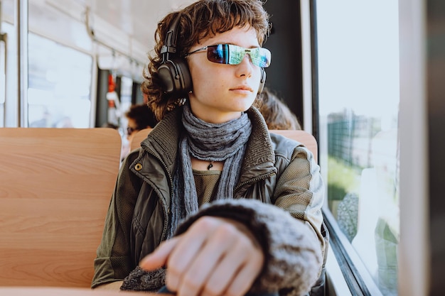 A boy wearing sunglasses and a jacket sits on a bus looking out a window