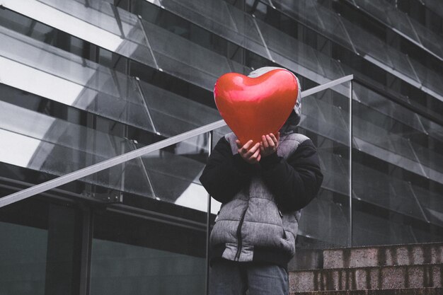 Photo boy wearing warm clothing while covering face with red heart shape balloon in city