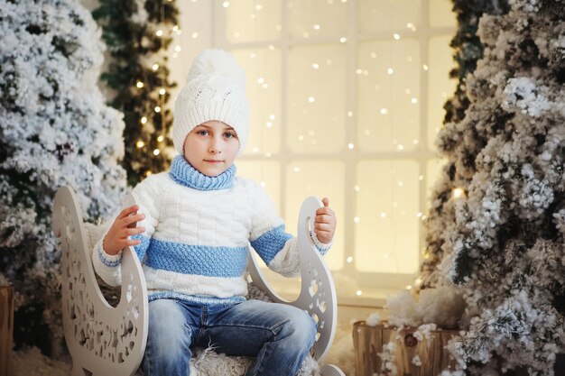 Boy in a white and blue sweater and white hat is sitting in a decorative sleigh near the Christmas tree.