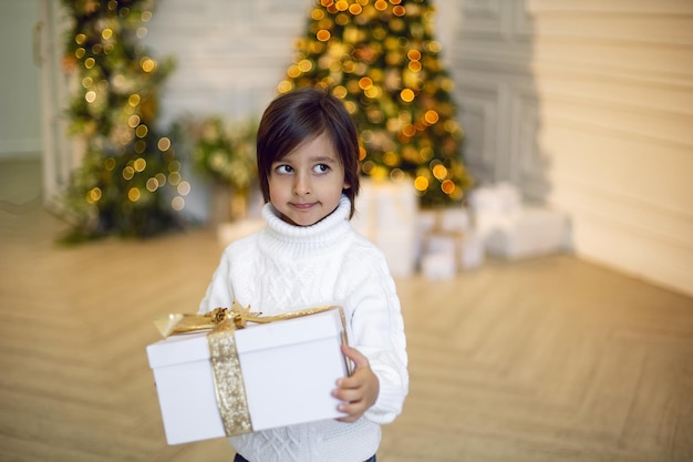 Boy in a white knitted sweater stands with a gift box at the Christmas tree at home on Christmas Day