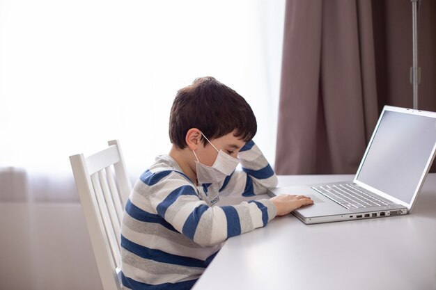 Un ragazzo con una maschera medica bianca, siede dietro un computer portatile