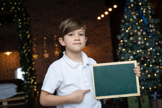 白いシャツを着た男の子がクリスマスツリーのそばに立って、テキスト用の空白のあるボードを持っています。クリスマス休暇のコンセプト