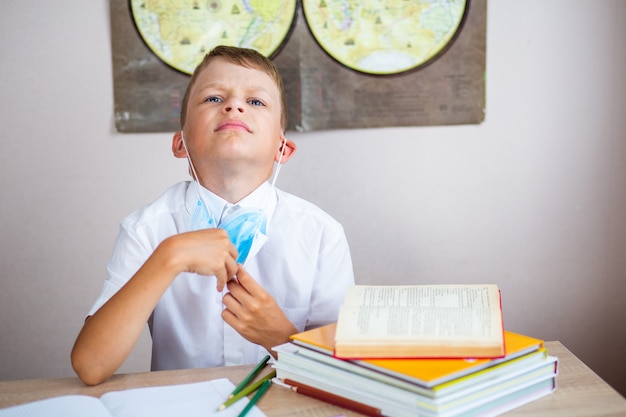 Boy in white shirt takes off medical mask sitting at his desk\
at school