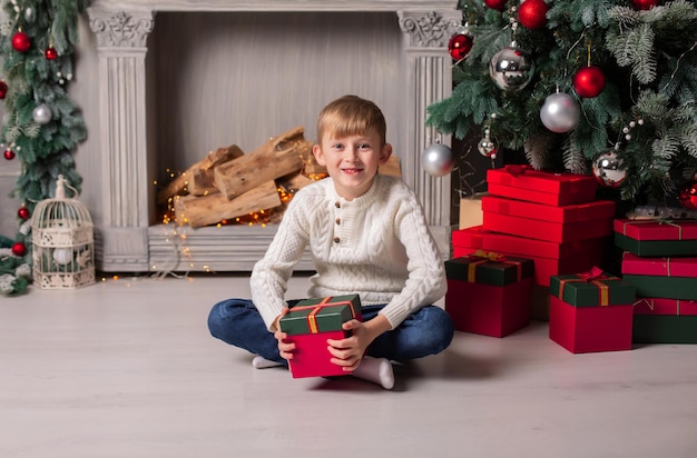 Un ragazzo con un maglione bianco è seduto accanto al camino di natale con una confezione regalo in mano