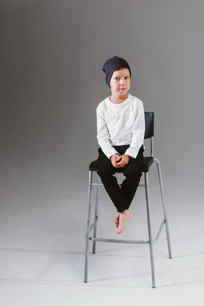 A boy in a white tshirt and cap sits on a bar stool on a white background Photoshoot of a boy model on a cyclorama