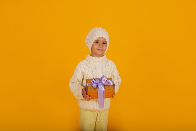 A boy in a white winter hat in a knitted white sweater holds a gift box in his hands