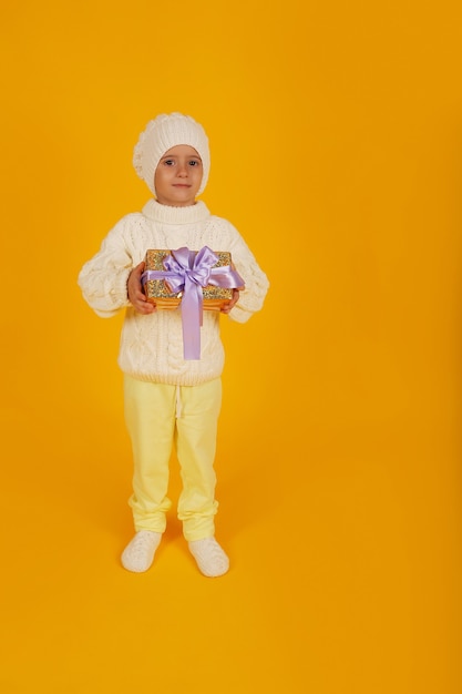 A boy in a white winter hat in a knitted white sweater holds a gift box in his hands