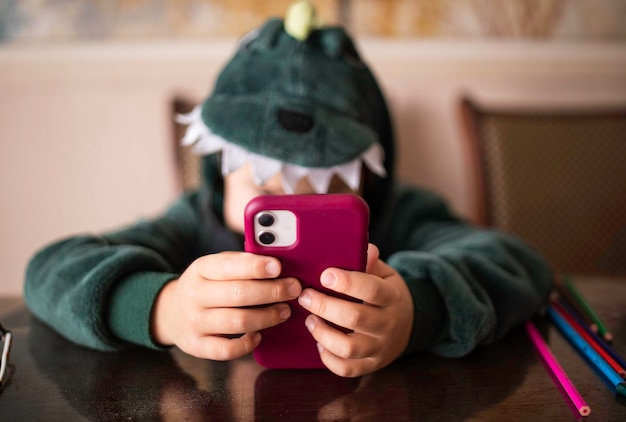 Boy who looks into the phone sitting in a dinosaur costume at the table unrecognizable face