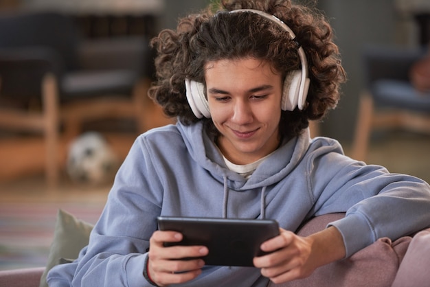 Photo boy in wireless headphones using his mobile phone for online conversation sitting at home