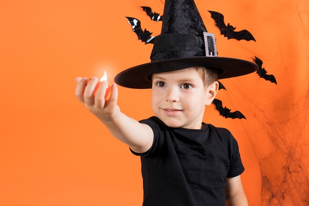 A boy in a witch costume in a black hat holds a small flashlight glowing pumpkin