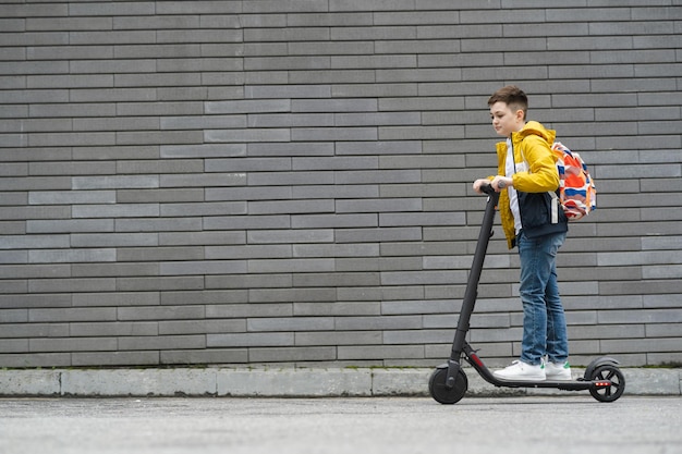 Boy with a backpack rides on electric scooter against a gray brick wall