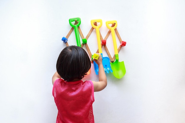 A boy with black hair aged 46 wearing a red tank top stands with his back playing with toys on a white wall with space to copy services or activities of baby supplies isolated on white background