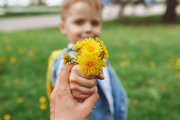 A boy with blond hair holds dandelions in his hands and holds them out to the camera against the backdrop of a green flowering field