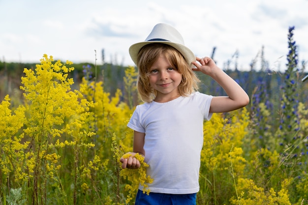 A boy with blond hair in a straw hat stands in a field with beautiful wild flowers