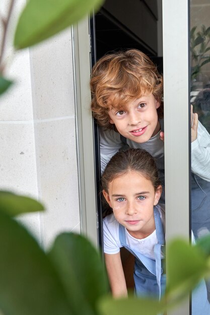 Boy with curly hair and girl in jeans overall looking out of glass doorway of the house