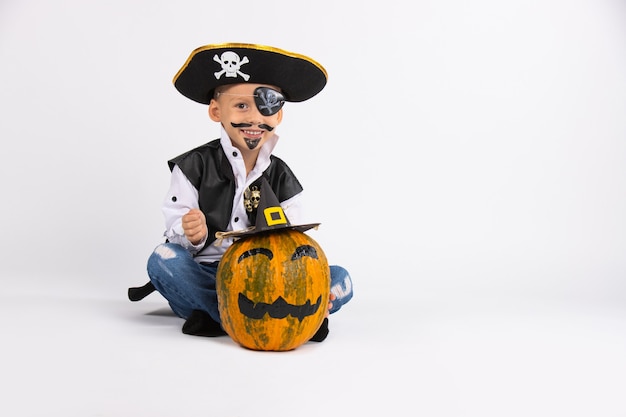 Boy with a cute smile wearing a pirate hat. Nearby is a pumpkin in a black handmade hat. Posing for the camera. 