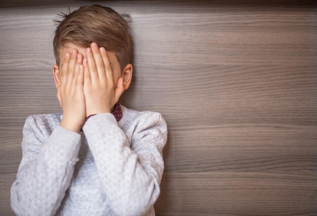 boy with face covered by hands against wooden wall
