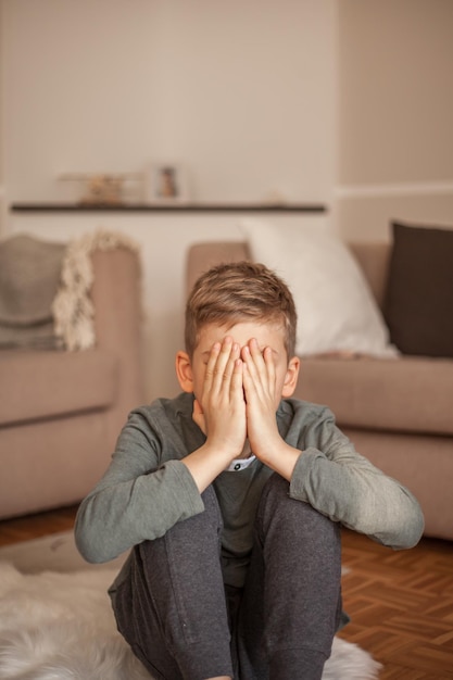 boy with face covered by hands sitting in living room at home