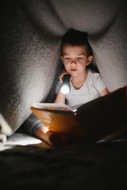 Boy with flashlight reading book under blanket at home