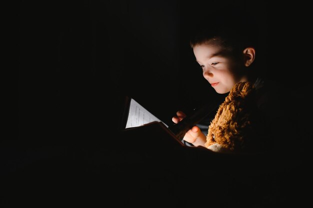 Boy with flashlight reading book under blanket at home