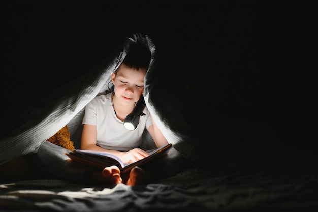 Boy with flashlight reading book under blanket at home