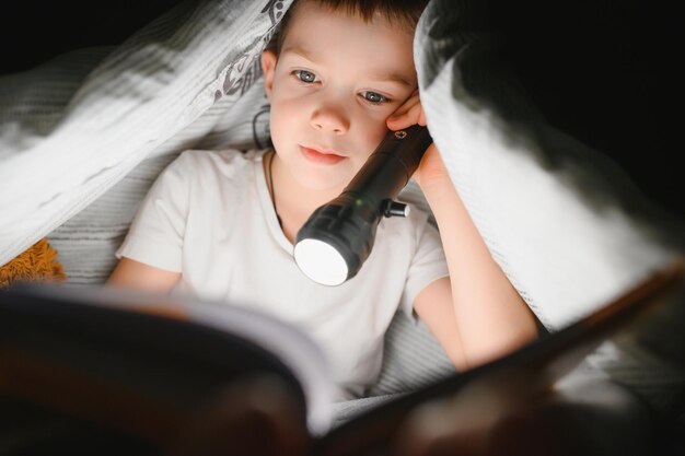 Boy with flashlight reading book under blanket at home