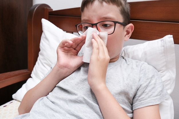 A boy with flu and fever lying in bed and blowing his nose with a paper tissue, seasonal viral diseases concept.