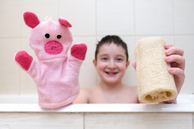 A boy with a funny face sitting in a bathtub and showing loofah and hand puppet washcloth close up.