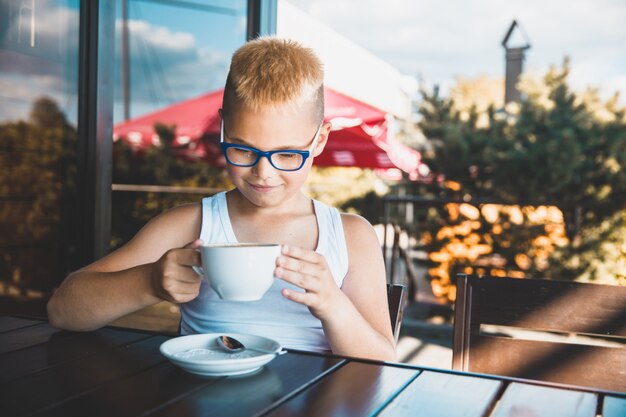 眼鏡をかけた少年がカフェに座ってコーヒーを飲む