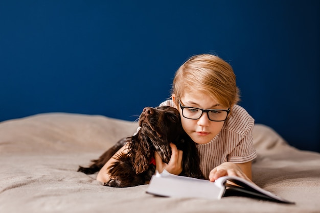 boy with glasses and with his dog is lying on the bed reading a big book.