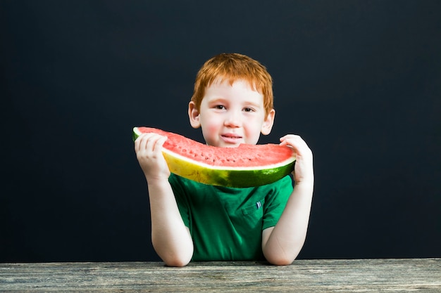 赤い髪の少年は、チャンクにカットされた赤いジューシーなスイカを食べる