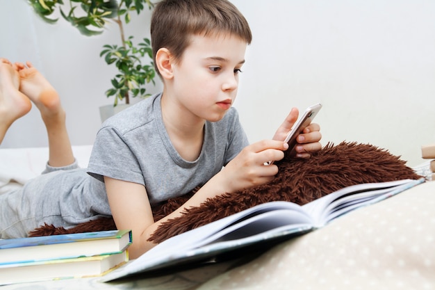 Мальчик с смартфоном в руках рядом с книгами в постели преподает уроки. Дистанционное обучение онлайн через Интернет