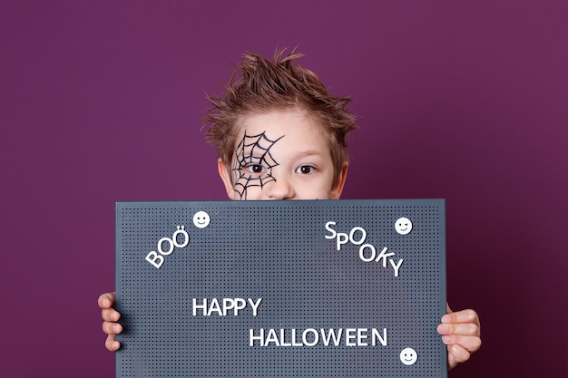 A boy with a spider web drawn on his face, holding a blackboard with the text of Halloween. Happy Halloween. Boo. Spooky.