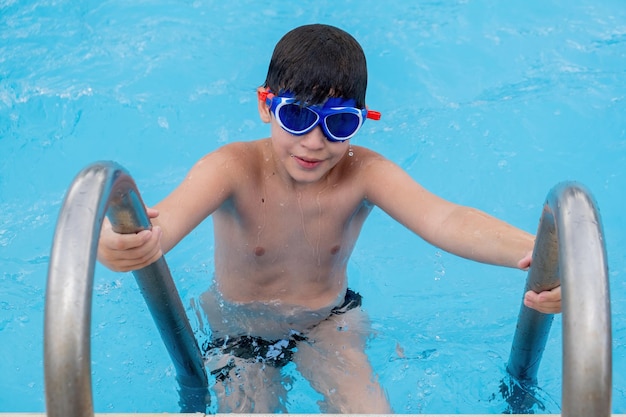 Photo boy with swimming googles leaves the pool from the ladder.