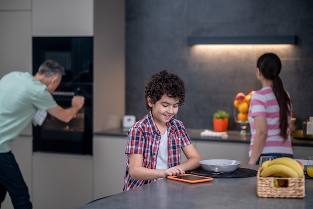 キッチンでの調理の背後にあるタブレットと両親を持つ少年