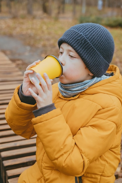 Boy in a yellow jacket drinks hot cocoa from a yellow glass sitting on a bench in the park the child