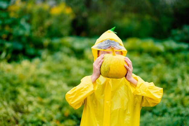 黄色のレインコートを着た少年が黄色いカボチャを持っている ハロウィーンの準備をしている カボチャの収穫 緑を背景に顔の近くの少年の手の中のカボチャ