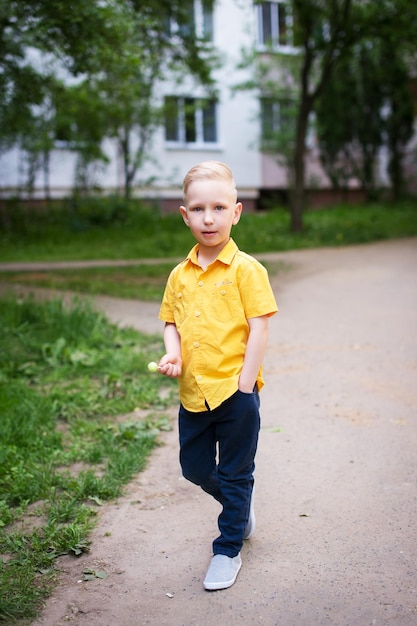 A boy in a yellow shirt and with white hair stands on the road and looks away