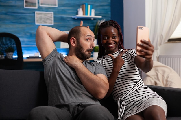 Boyfriend being goofy while joyful girlfriend taking selfie using smartphone. Cheerful african american wife and caucasian husband taking pictures together with phone while sitting on sofa in living r