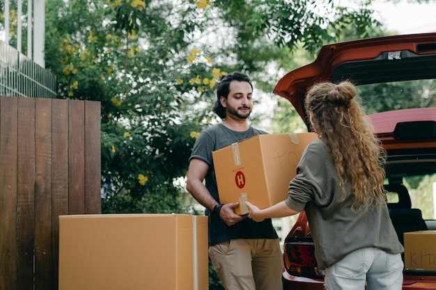 Photo boyfriend and girlfriend in casual wear helping each other with unpacking car while moving in togeth