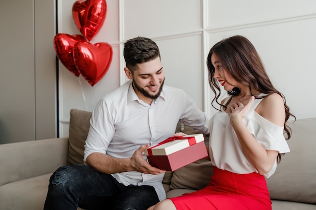 Boyfriend giving gift in present box to girlfriend with heart shaped balloons on couch at home