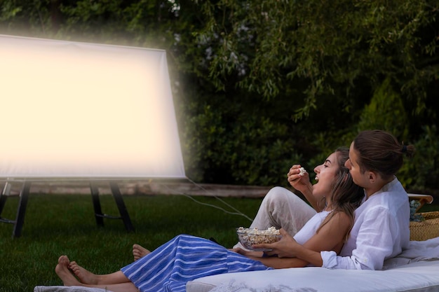 Photo boyfriend giving popcorns to his girlfriend in the open mouth as a sign of love while in a cinema date in summer