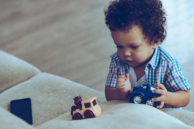 Boys love little trains. Little African baby boy playing with toys while standing near the couch at home