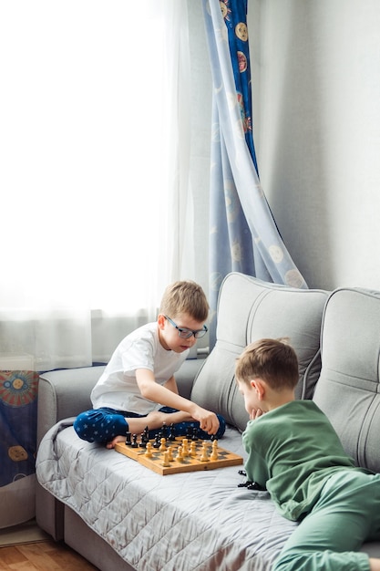 Boys playing wooden chess lying on a gray sofa in front of the window